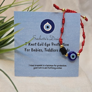 May include: A red string bracelet with gold beads and a blue evil eye charm. The bracelet is displayed on a light blue card with text that reads "7 Knot Evil Eye Protection For Babies, Toddlers & Kids."