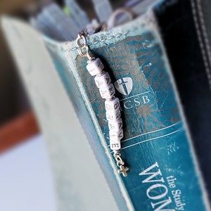 May include: A white beaded bookmark with the words "LOVE" and "GRACE" on it. The bookmark is attached to a blue book with the words "the Study Bible" and "CSB" on the cover.
