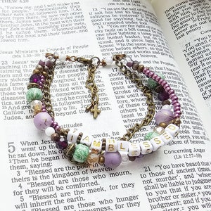 May include: A multi-strand bracelet with beads in shades of purple, green, and white, accented with gold-toned metal and a small cross charm. The bracelet spells out "BLESSED" with white letter beads. The background is a Bible.
