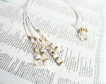 May include: A handmade bookmark with a white cord, adorned with gold-colored beads and white, pearlescent beads. The bookmark is placed on an open book with text.