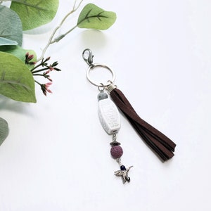 May include: White ceramic charm with a floral design, a gray bead, a purple lava rock bead, and a silver hummingbird charm attached to a brown suede tassel and a silver key ring.