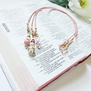 May include: A pink and white beaded lanyard with a gold ring and charms. The lanyard is laying on a page of an open book with text about the Lord.