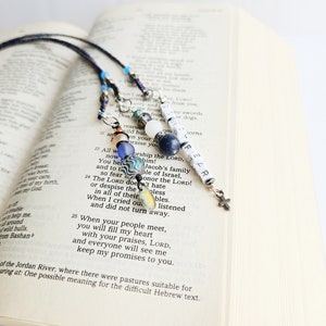 May include: Three blue, white, and black beaded bookmarks with silver charms and a small cross charm. The bookmarks are hanging from a Bible open to a page with text about the Lord.