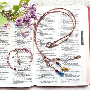 May include: A necklace and bracelet set with the word "WORTHY" spelled out in white beads. The jewelry is displayed on an open book with purple flowers.