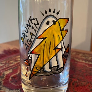 Könnte beinhalten: Ein klares Glas mit einem handgemalten Design eines Geistes, der einen Blitz hält. Der Geist trägt einen gelben Umhang und der Text "DRUNK AGAIN" ist auf das Glas geschrieben.