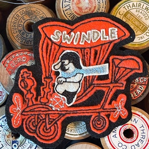 May include: A black and orange embroidered patch with the word "SWINDLE" in white lettering. The patch features a cartoon character driving a vehicle with wheels.