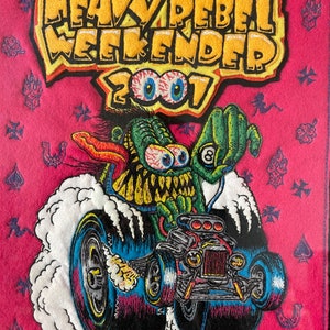 May include: A pink felt patch with a yellow embroidered patch that says "Heavy Rebel Weekender 2001". A green monster with large eyes is driving a hot rod with flames coming out of the back tires.
