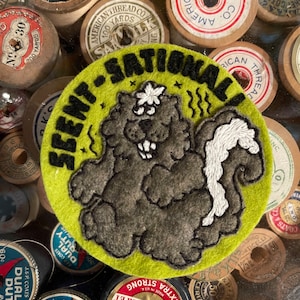 May include: A green and black embroidered patch with a cartoon skunk with a white tail and the text "Scent-sational" in black.