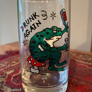Könnte beinhalten: Ein klares Glas mit einem handgemalten Design, das einen grünen Frosch zeigt, der eine Flasche und ein Glas hält. Die Worte "DRUNK AGAIN" sind auf dem Glas geschrieben. Der Frosch sitzt auf einem roten und weißen Pilz.