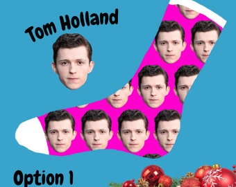 Peut inclure: Une paire de chaussettes roses avec un bout et un talon blancs. Les chaussettes présentent un motif répété d'un visage d'homme. Le texte "Tom Holland" est écrit au-dessus des chaussettes. Les chaussettes sont posées sur un fond bleu avec une décoration de Noël dans le coin inférieur droit.