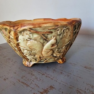 May include: A ceramic bowl featuring a raised relief design of a squirrel perched on tree branches. The bowl has a scalloped rim, three small feet, and a glossy glaze in shades of brown, tan, and muted green, creating a textured appearance.