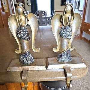 Könnte beinhalten: Zwei goldfarbene Engel-Strumpfhalter. Jeder Engel hat ausgebreitete Flügel, einen Heiligenschein und einen dekorativen Rock mit Mosaikdesign. Die Halter haben einen Haken zum Aufhängen von Strümpfen.