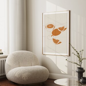 Peut inclure: Une impression d'art minimaliste repr&eacute;sentant un verre de jus d'orange avec des tranches d'orange sur un fond beige. L'impression est encadr&eacute;e dans un cadre en bois clair et est accroch&eacute;e &agrave; un mur blanc.