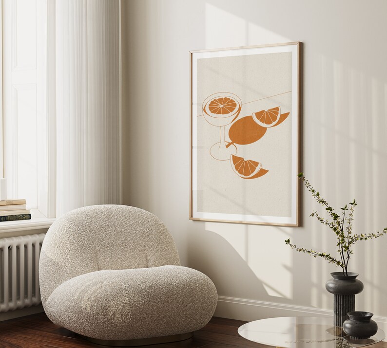 Peut inclure: Une impression d'art minimaliste repr&eacute;sentant un verre de jus d'orange avec des tranches d'orange sur un fond beige. L'impression est encadr&eacute;e dans un cadre en bois clair et est accroch&eacute;e &agrave; un mur blanc.