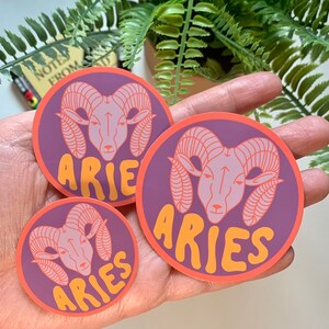Puede incluir: Tres pegatinas redondas de color morado y naranja con una cabeza de carnero estilizada y la palabra "Aries" en una fuente redondeada.
