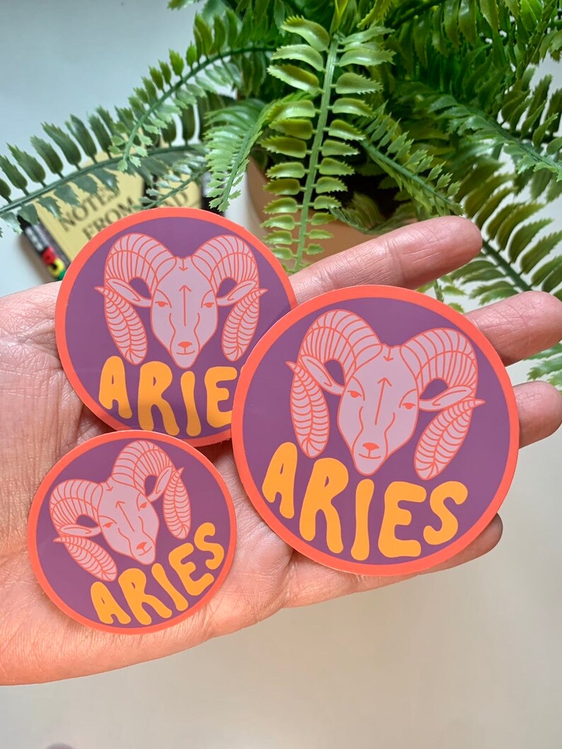 Puede incluir: Tres pegatinas redondas de color morado y naranja con una cabeza de carnero estilizada y la palabra "Aries" en una fuente redondeada.