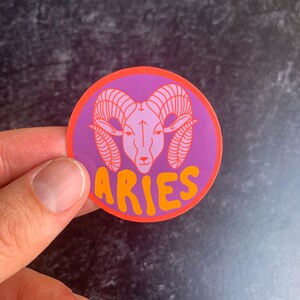 Puede incluir: Una pegatina redonda con fondo morado y borde naranja. La pegatina presenta una cabeza de carnero rosa con cuernos naranjas y la palabra "ARIES" en letras naranjas.