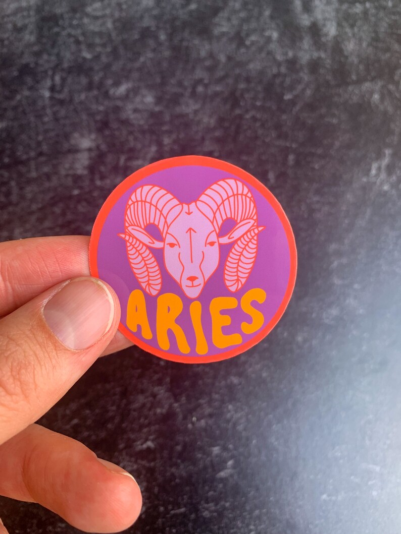 Puede incluir: Una pegatina redonda con fondo morado y borde naranja. La pegatina presenta una cabeza de carnero rosa con cuernos naranjas y la palabra "ARIES" en letras naranjas.