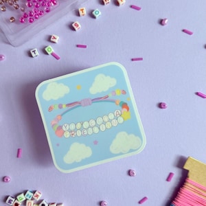 May include: A square light blue sticker featuring a graphic of a beaded bracelet with the text YOU'VE GOT A FRIEND IN ME, surrounded by scattered alphabet beads, a clear plastic bead organizer, and a spool of pink, blue, and purple thread.