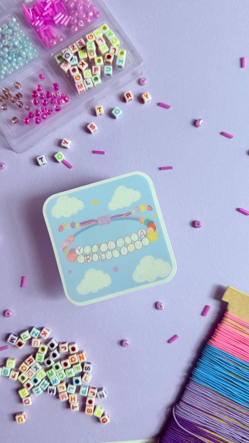 May include: A square light blue sticker featuring a graphic of a beaded bracelet with the text YOU'VE GOT A FRIEND IN ME, surrounded by scattered alphabet beads, a clear plastic bead organizer, and a spool of pink, blue, and purple thread.
