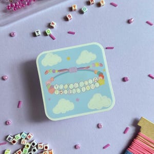May include: A square sticker featuring a light blue sky with white clouds and a colorful beaded bracelet illustration. The bracelet has letter beads spelling "YOU'VE GOT A FRIEND IN ME" and is surrounded by loose alphabet beads and colorful string.