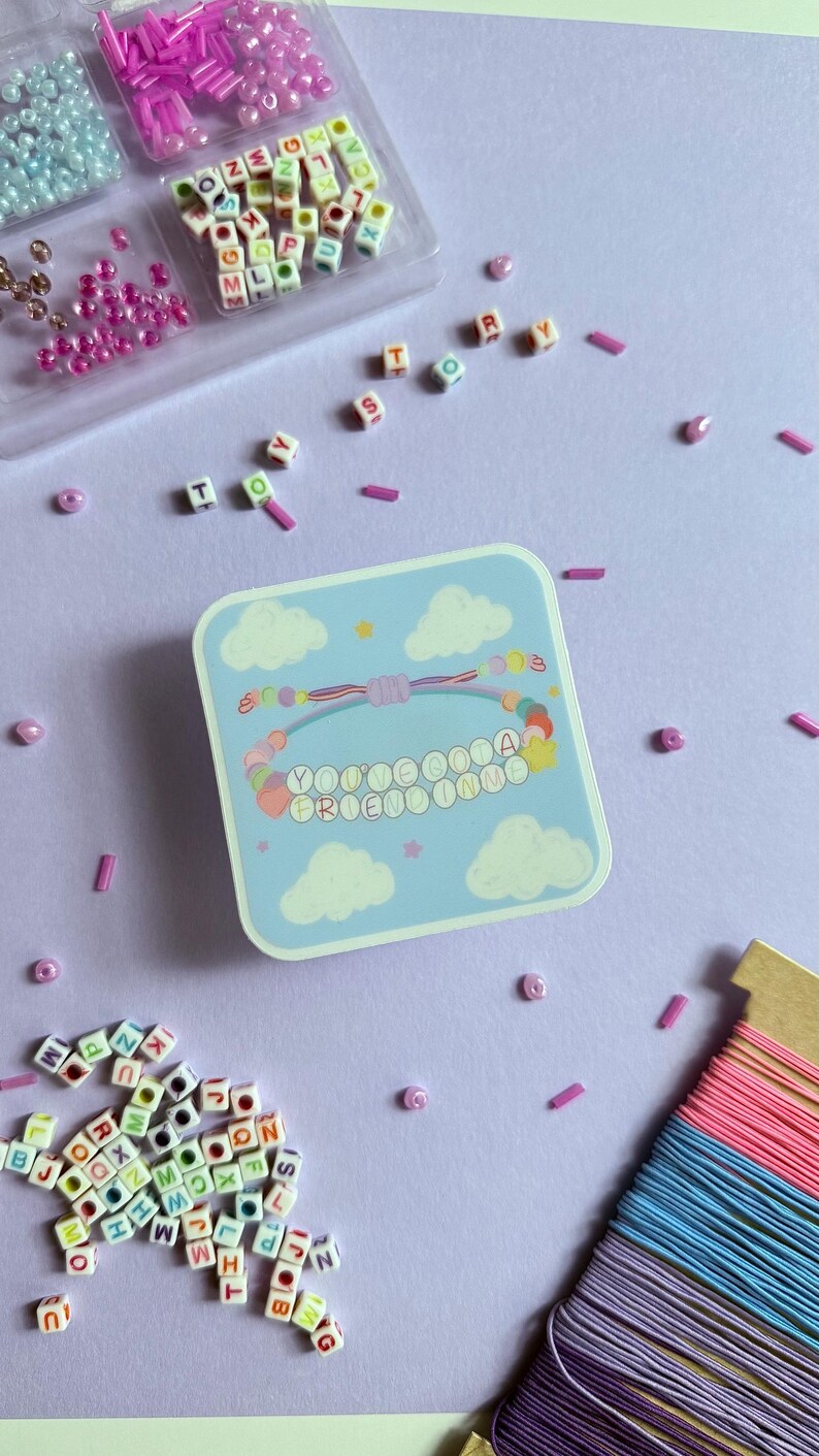 May include: A square sticker featuring a light blue sky with white clouds and a colorful beaded bracelet illustration. The bracelet has letter beads spelling "YOU'VE GOT A FRIEND IN ME" and is surrounded by loose alphabet beads and colorful string.