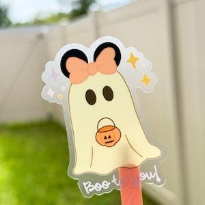 May include: A clear sticker featuring a cartoon ghost with a peach bow, black ears, and a jack-o'-lantern. The ghost is holding a pumpkin bucket and has the text "Boo to you!" in a script font. The background is blurred.