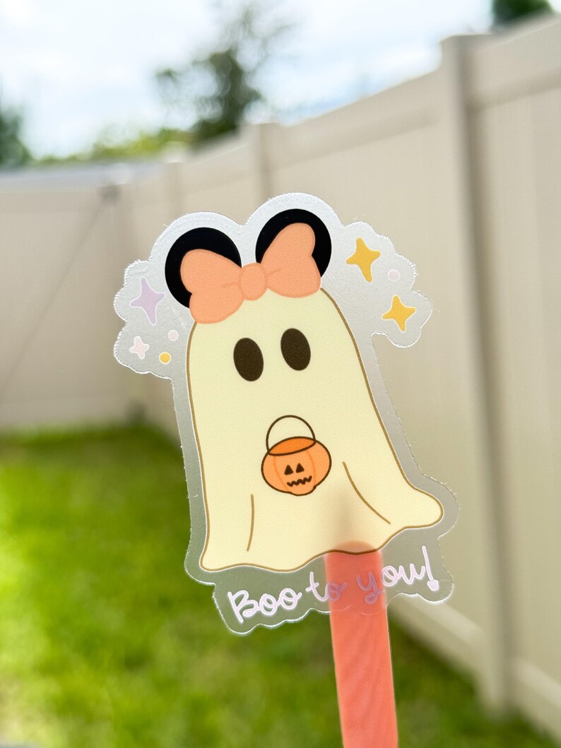 May include: A clear sticker featuring a cartoon ghost with a peach bow, black ears, and a jack-o'-lantern. The ghost is holding a pumpkin bucket and has the text "Boo to you!" in a script font. The background is blurred.