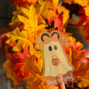 May include: A Halloween-themed pen with a ghost sticker that says "Boo to you!" The ghost has a bow and is holding a pumpkin bucket. The background is a wreath of orange and yellow fall leaves.