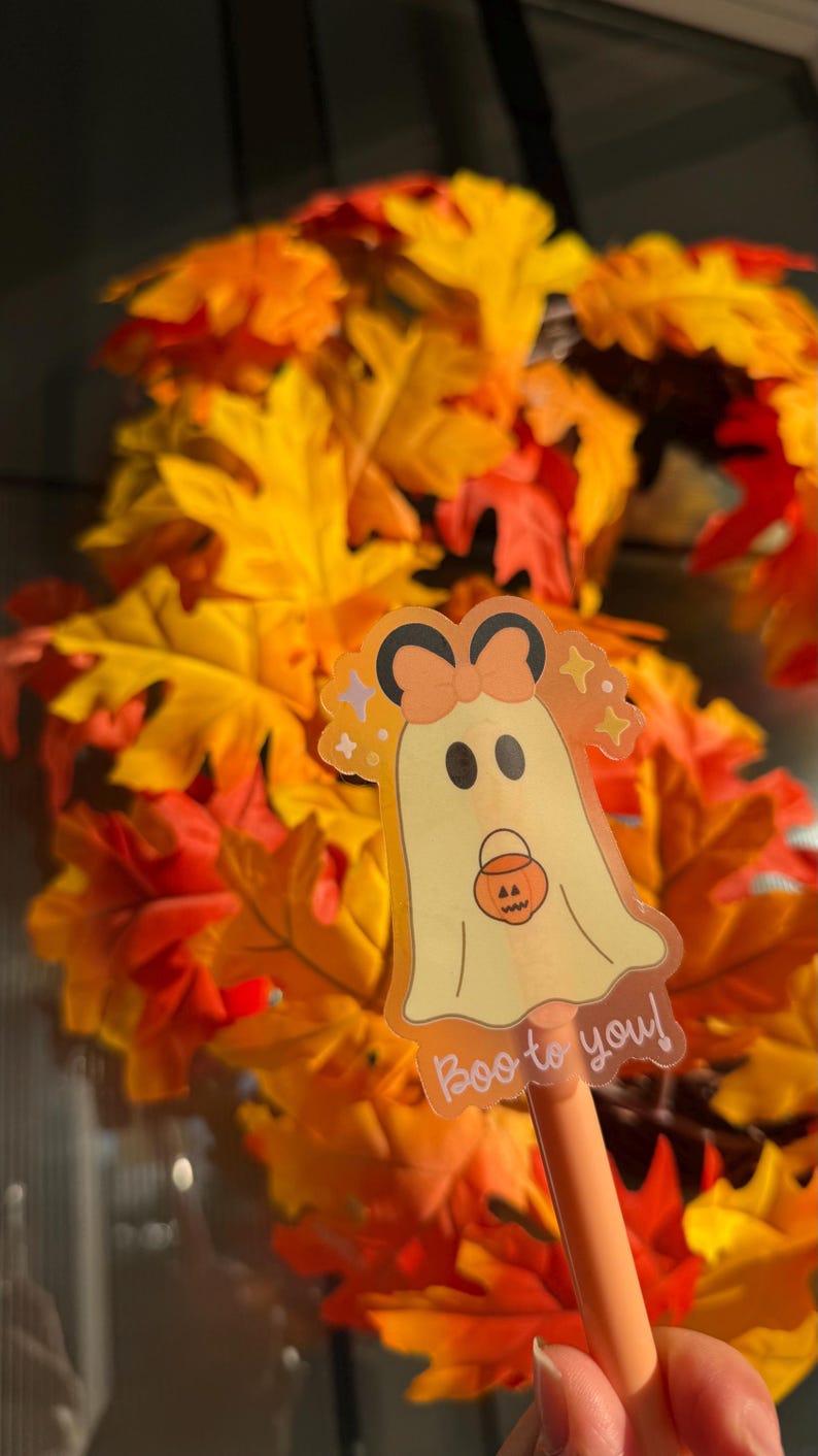 May include: A Halloween-themed pen with a ghost sticker that says "Boo to you!" The ghost has a bow and is holding a pumpkin bucket. The background is a wreath of orange and yellow fall leaves.