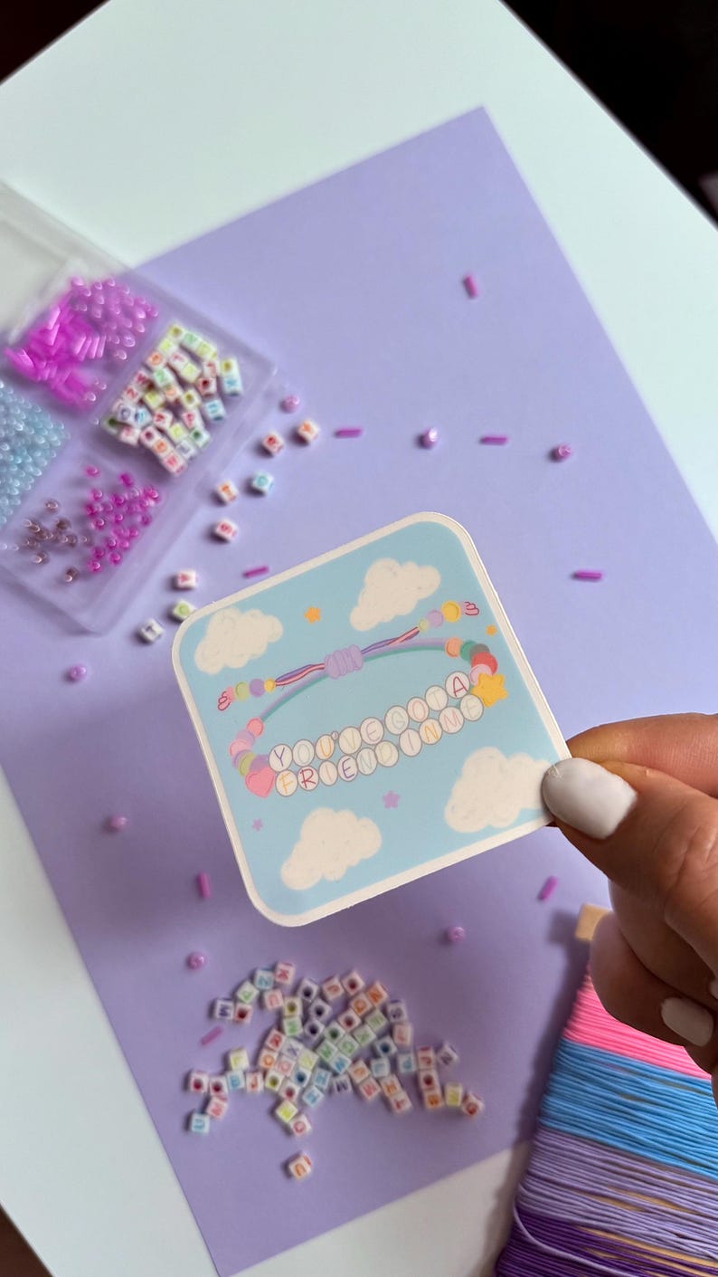 May include: A square sticker featuring a light blue sky background with white clouds and a colorful beaded bracelet illustration. The bracelet has letter beads spelling out the phrase YOU'VE GOT A FRIEND IN ME, accented with small hearts and stars.