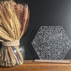 Könnte beinhalten: Ein sechseckiges, klares Acrylschild mit einem weißen Blumenmuster. Das Schild wird von hinten beleuchtet und steht auf einem Holzsockel.