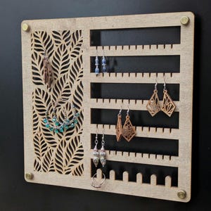 May include: A wooden earring display board with a leaf pattern cut-out. The board features multiple horizontal slots for hanging earrings. Various earrings of different styles and colours are displayed on the board.