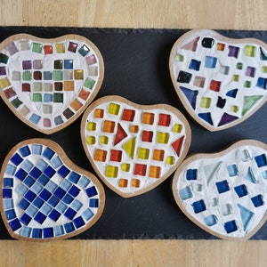 May include: Five heart-shaped coasters made of wood and decorated with colourful mosaic tiles. The coasters are arranged in a row on a black slate surface.