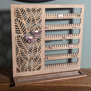 May include: Wooden earring holder with a leaf design and multiple rows of holes for earrings. The holder has a brown finish and a small base.