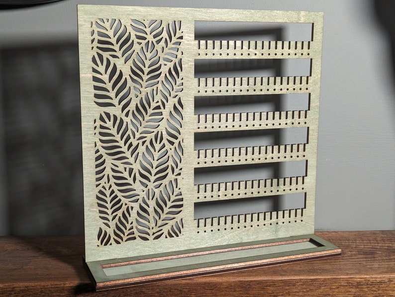 Puede incluir: Organizador de pendientes de madera con un dise&ntilde;o de patr&oacute;n de hojas y m&uacute;ltiples filas de agujeros para guardar pendientes.
