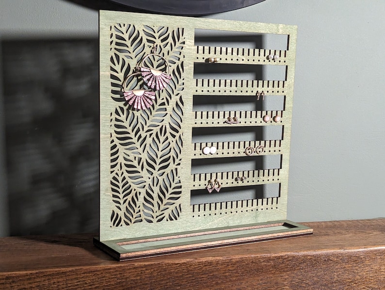 Puede incluir: Un soporte de madera para pendientes con un dise&ntilde;o de hojas y varias filas de agujeros para pendientes. El soporte tiene una peque&ntilde;a repisa en la parte inferior.