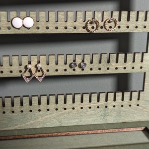 Puede incluir: Un soporte de madera para pendientes con m&uacute;ltiples filas de agujeros para guardar pendientes. El soporte est&aacute; pintado de color verde claro y tiene un peque&ntilde;o caj&oacute;n en la parte inferior.