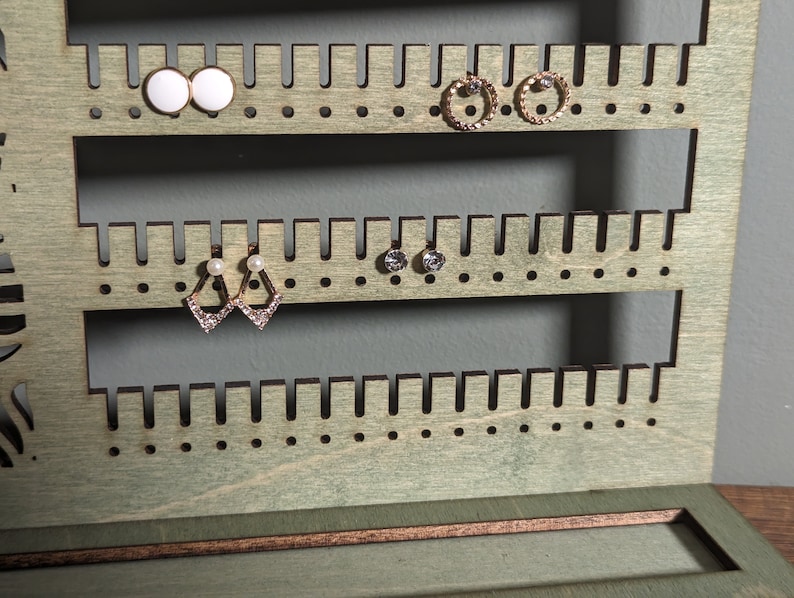 Puede incluir: Un soporte de madera para pendientes con m&uacute;ltiples filas de agujeros para guardar pendientes. El soporte est&aacute; pintado de color verde claro y tiene un peque&ntilde;o caj&oacute;n en la parte inferior.