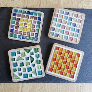 Könnte beinhalten: Vier quadratische Mosaik-Untersetzer mit Holzrahmen. Jeder Untersetzer hat ein einzigartiges Design aus farbigen Glasfliesen. Die Untersetzer sind auf einer dunkelgrauen Schieferoberfläche angeordnet.