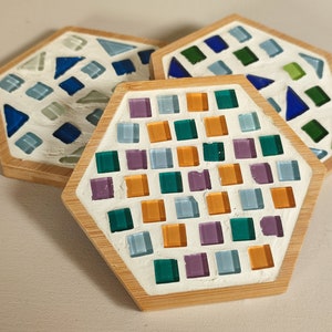 Könnte beinhalten: Drei sechseckige Untersetzer aus Holz mit einer weißen Basis und bunten Glasmosaikfliesen. Die Untersetzer zeigen verschiedene Muster aus blauen, grünen, orangefarbenen und violetten Fliesen.