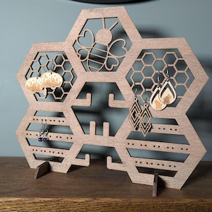 May include: Wooden honeycomb-shaped earring holder with a bee design in the centre. The holder has multiple slots and pegs for storing earrings.