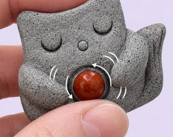 May include: A gray, textured, stone-like cat figurine with closed eyes, holding a smooth, round, polished red gemstone in its center. White curved lines are drawn around the red stone to indicate a spinning or rotating motion of the object.