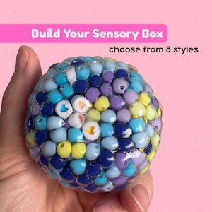 May include: A round, clear sensory stress ball filled with a dense cluster of small, multi-colored plastic beads in shades of blue, purple, yellow, and white. Some white beads feature small heart-shaped cutouts. Text reads: Build Your Sensory Box choose from 8 styles.
