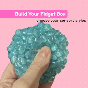 May include: A round, turquoise, glittery sensory fidget toy composed of numerous small, translucent spheres embedded in a clear gel. The text 'Build Your Fidget Box' and 'choose your sensory styles' is displayed on a pink background above the toy.