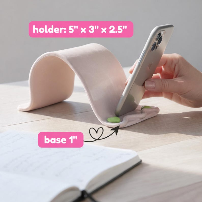 May include: A light pink phone holder with a curved design, dimensions 5" x 3" x 2.5". The base is 1" thick. A smartphone rests in the holder. Two small green pads are visible. A notebook is in the foreground.