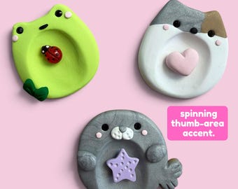 May include: Three small, round, sculpted trinket dishes featuring animal designs: a lime green frog with a ladybug, a white and gray cat with a pink heart, and a gray seal with a purple star. A pink label reads: spinning thumb-area accent.