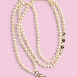 May include: A cream-colored pearl beaded lanyard with the name "JANINE" spelled out in gold letter beads. The lanyard has a silver-colored clasp and a pink background.