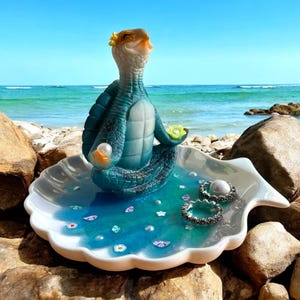 May include: A decorative jewelry dish featuring a meditating turtle figurine. The turtle is teal and brown, seated in a shell-shaped dish with a blue and turquoise water effect. The dish holds rings and decorative elements.