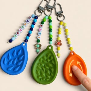 May include: Three keychains with colorful beaded strands and teardrop-shaped silicone charms. One blue charm has the name "CAMERON" spelled out in beads. The other keychains have green and orange charms. The green charm has a textured surface.